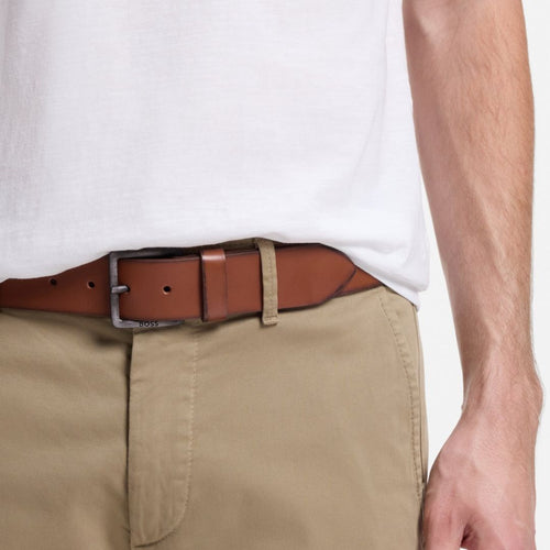 A close-up of a person wearing a brown BOSS leather belt with a silver-toned buckle, tucked into khaki pants and a white t-shirt.