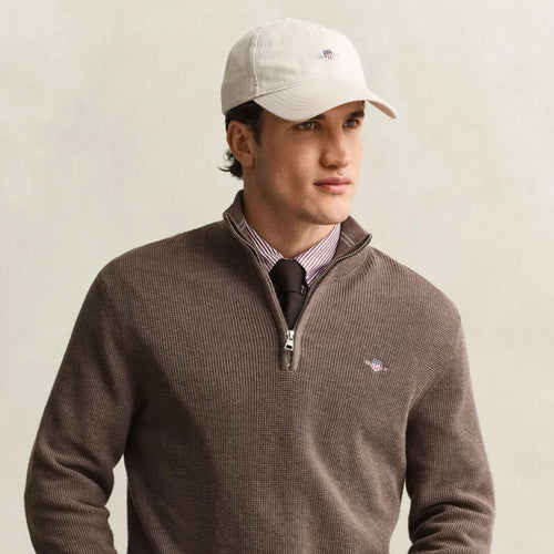 A man wearing a brown ribbed quarter-zip sweater over a striped collared shirt and black tie, paired with a light beige Gant baseball cap. The sweater features a small embroidered logo on the left chest.