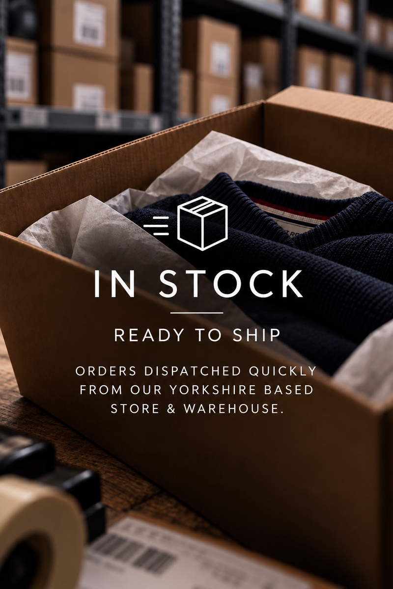 A brown cardboard shipping box filled with a dark navy knit garment and tissue paper, with overlaid text reading 'IN STOCK READY TO SHIP ORDERS DISPATCHED QUICKLY FROM OUR UK WAREHOUSE.' The background shows blurred warehouse shelves and boxes.